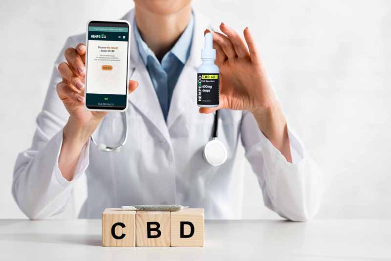 Doctor with smart phone and Hempexco CBD website showing on the screen. There is a bottle of Hempexco CBD in her other hand
